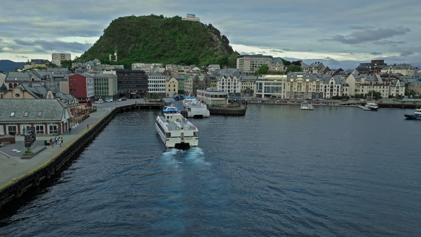 Alesund, Norway aerial drone view centered on boats navigating toward the harbour basin.The norwegian scene shows multiple vessels, curved piers, dense buildings along the shoreline, and a green hill rising behind the town.