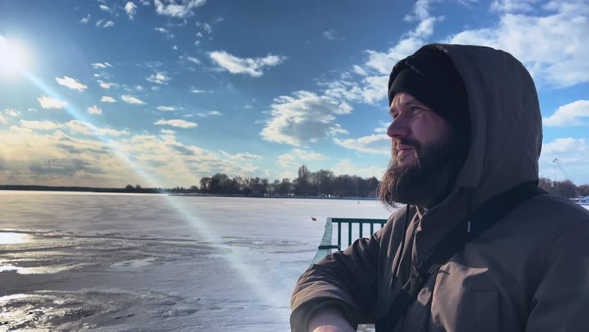A stern, bearded man stands on the shore of a frozen lake and looks into the distance.