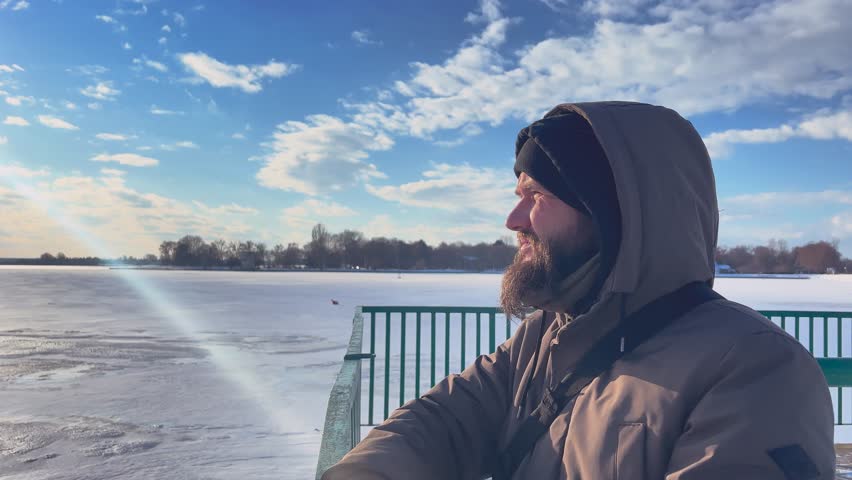 A stern, bearded man stands on the shore of a frozen lake and looks into the distance.
