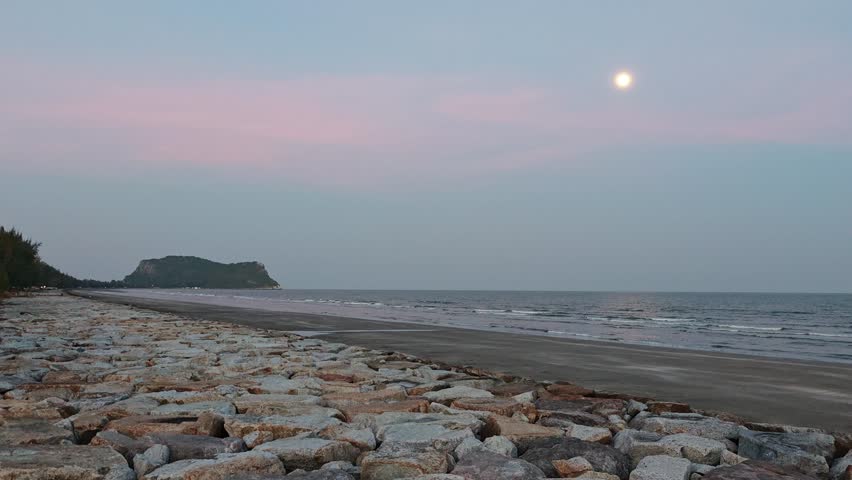 Beautiful rocky coastline with calm sea waves and moon in the sky. Peaceful stone beach landscape during twilight evening.