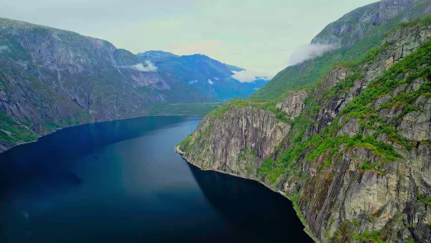 Aerial view of a serene Norwegian fjord, showcasing dramatic cliffs plunging into dark, reflective waters under a cloudy sky