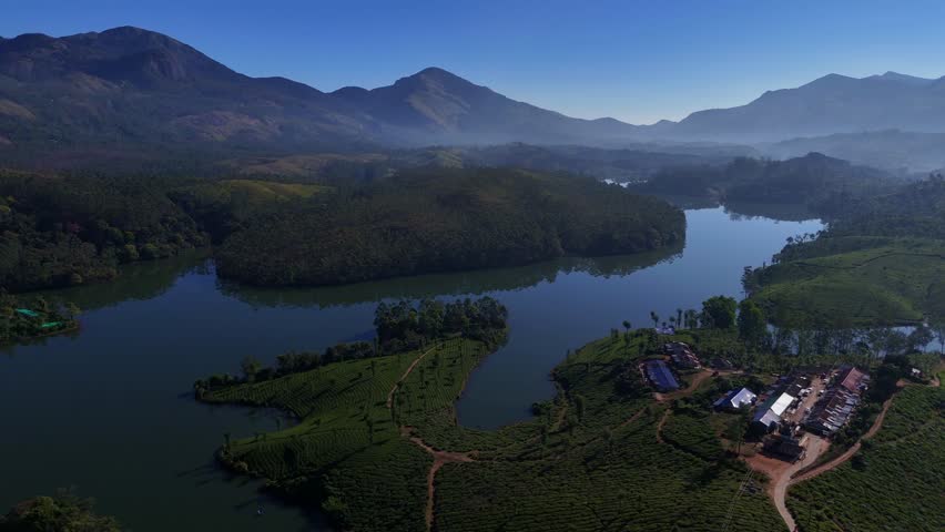 Cinematic aerial drone flight over the lush tea plantations of Munnar, Kerala, India, revealing winding roads disappearing into soft morning mist and serene lakes far below. The layered green hills and drifting fog create a tranquil atmosphere, capturing the beauty of South India’s highland landscape at sunrise. אידeal for themes of travel, eco tourism, nature, agriculture, serenity, and scenic destinations.