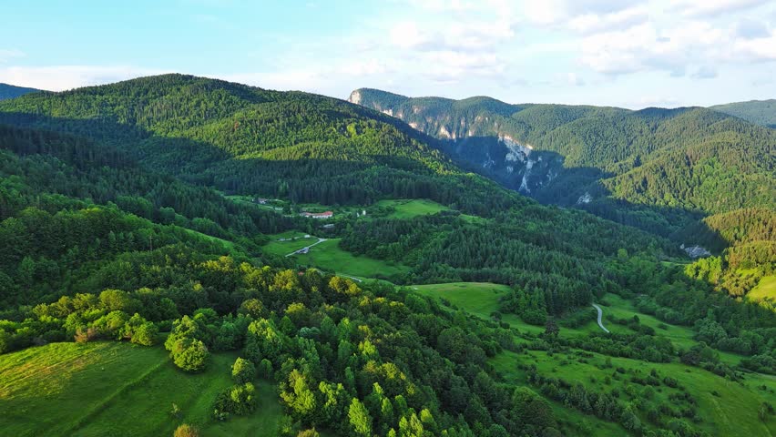 A road passes hills covered with spruce forests and meadows against a cloudy sky
