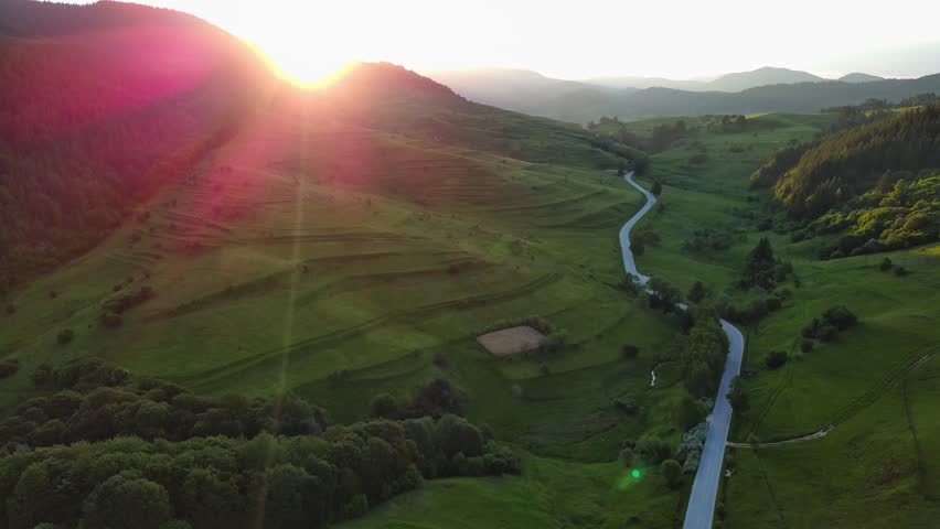 A road passes hills covered with spruce forests and meadows against a cloudy sky