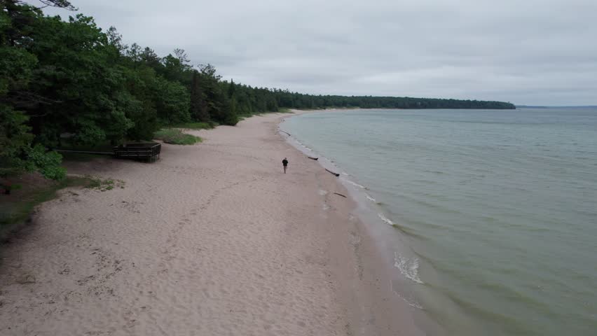 Follow drone shot of man on beach in Northern Door County Wisconsin.  Lake Michigan water a tree lined shore.