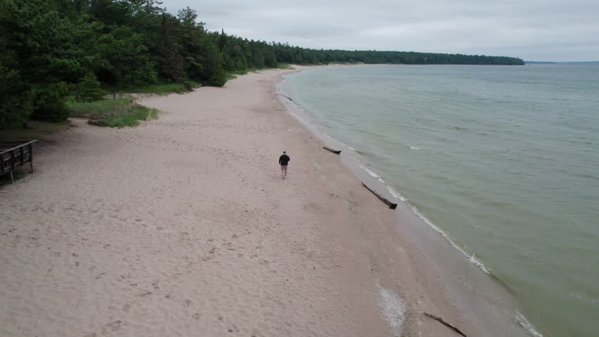Follow drone shot of man on beach in Northern Door County Wisconsin.  Lake Michigan water a tree lined shore.