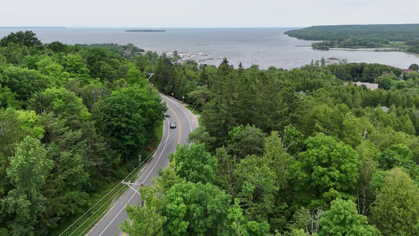 Aerial of Fish Creek in Door County Wisconsin on an overcast day. Drone push shot over road and bluff with cars and hilly terrain in the distance.  Island and harbor on the horizon. 