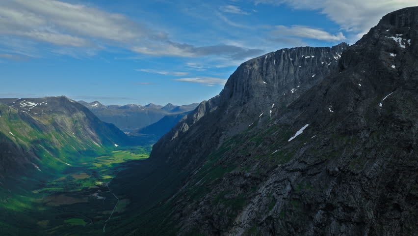 Drone view opens to a wide valley at Trollstigen framed by towering cliffs and green lowlands. Beyond Trollstigen’s rock walls, distant hills and layered terrain complete the scene.
