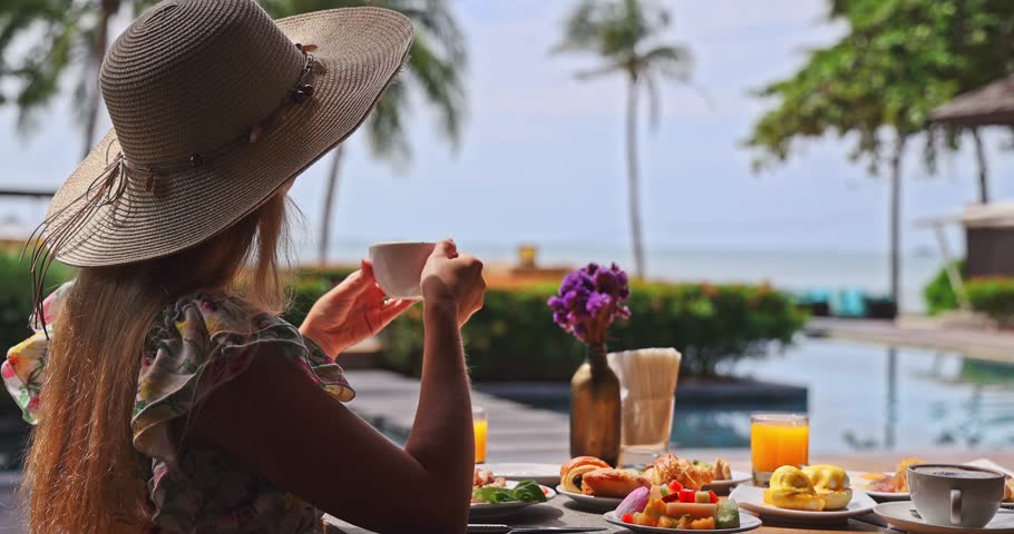 Woman wearing straw hat drinking coffee at luxury tropical resort breakfast table, poolside ocean view with palm trees, bright morning light, relaxing vacation lifestyle, hospitality travel.