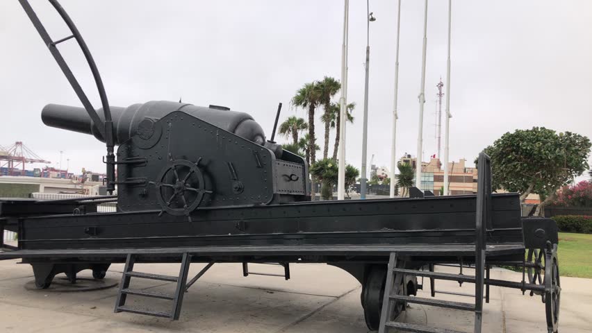 historic naval cannon on trailer callao, black armored gun mounted on flatbed near harbor cranes and palms, overcast sky, concrete plaza, riveted metal turret, museum