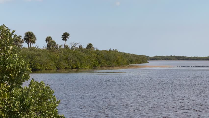 Rocket launch pad at Space Center in Florida is seen across a calm lagoon under a clear blue sky. Communication towers, a water tower, and the green shoreline frame the space exploration facility