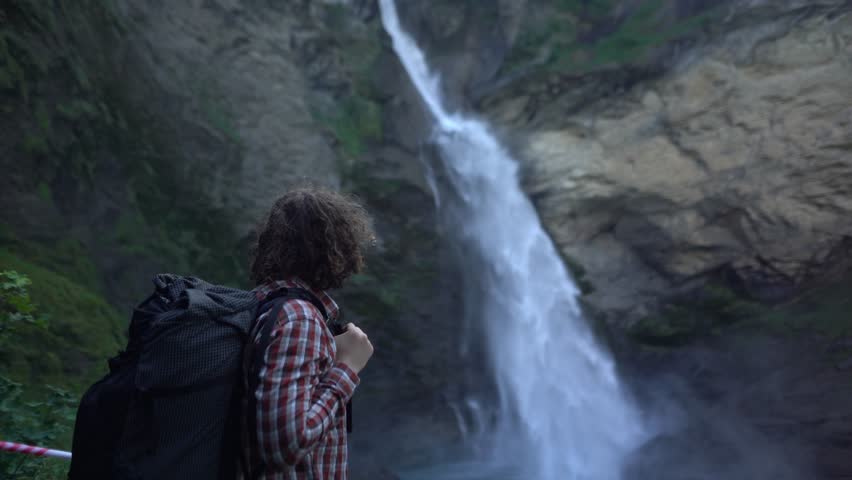Backpacker hiking close to a powerful alpine waterfall in the Swiss Alps, pausing to observe flowing water and steep rock walls. Reichenbach waterfall near Meiringen, dramatic nature outdoor exploring