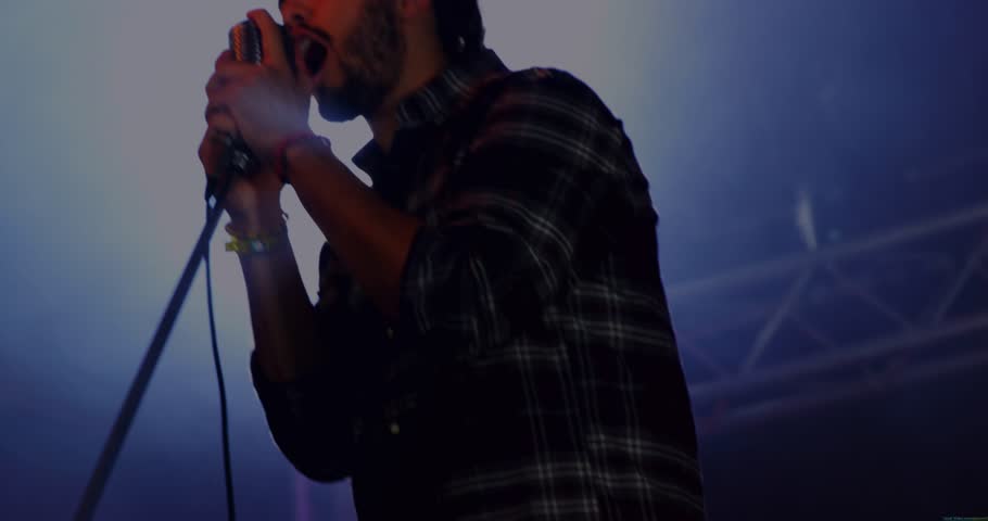 Male singer gripping vintage mic and performing song on music stage backlight flaring into shadow. Silhouette, lensflare, backlit, haze, concert, lights, plaid