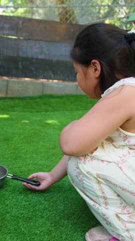 A young girl crouches on green turf, offering food to a rabbit in a pan
