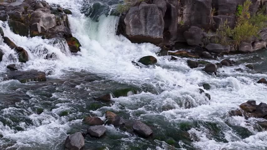 Drone footage captures a small waterfall and rapids in the Snake River at Idaho Falls, with clear water cascading over dark rocks and forming frothy whitewater against rocky riverbanks.
