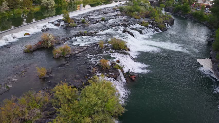 Aerial drone footage captures the Snake River waterfalls with water cascading over rocks and islands, creating whitewater rapids surrounded by green vegetation and trees