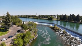 Drone view of the Snake River at Idaho Falls, with water flowing over a curved dam and weir beside a tree-lined riverbank and road with cars, park areas and city skyline visible. - Powered by Shutterstock - Get 15% off with code: PIKWIZARD15