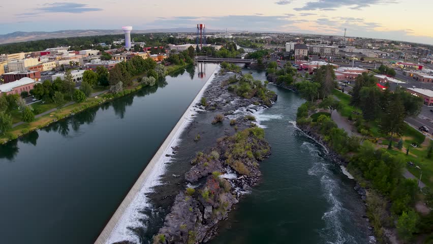 Aerial footage of Snake River weir with walkway and cascading water, surrounded by park and city skyline near Idaho Falls in Idaho