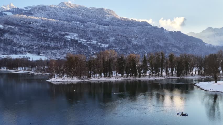 Winter shoreline of Walensee with snowy trees, calm water reflections, and alpine mountains in the background.