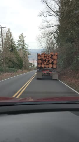 Logging truck transporting cut timber driving on a two lane forest road in Oregon, USA, Pacific Northwest. Rear view video footage of a loaded log truck moving through wooded rural landscape and a winding highway. Real world transportation footage representing forestry industry, timber harvest, natural resources, and regional cargo transport.