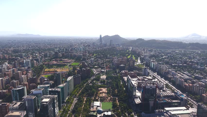 Wide aerial drone shot capturing the vastness of Santiago, Chile, from Parque Araucano to the distant Costanera Center, illustrating the blend of urban parks and dense metropolitan infrastructure.