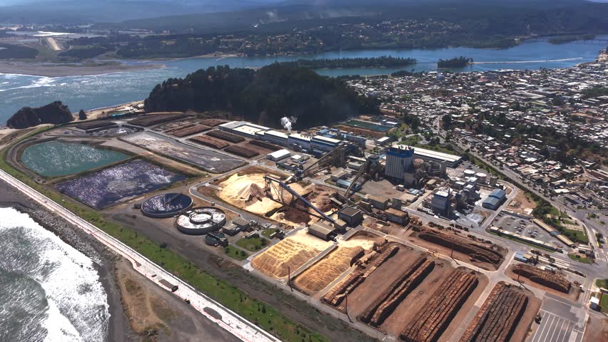 Wide aerial angle of a massive industrial pulp mill complex located on the coastline near the river mouth in Constitución, Chile, showing wood stockpiles and factory layout.