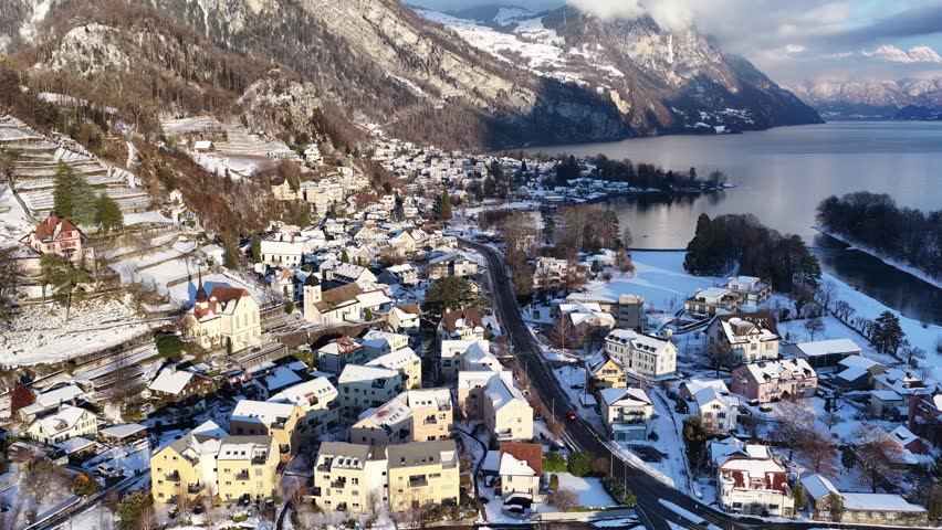 Panoramic drone view of Weesen, Switzerland, in winter. Snowy roofs, the historic church, and the calm lake harbor are nestled beneath the majestic, hazy Alpine peaks.