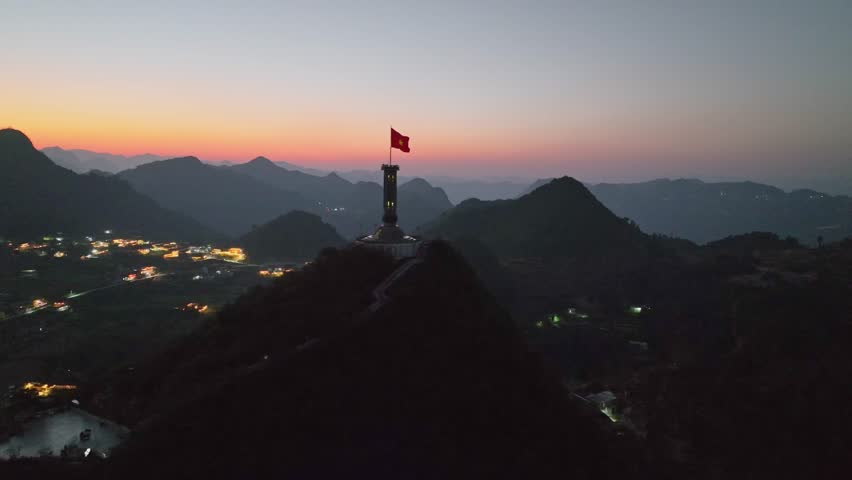 Twilight view of Lung Cu Flag Tower silhouetted on mountain peak with glowing village lights below.