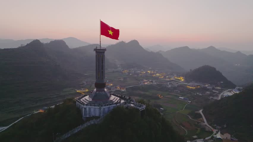 Aerial twilight view of Lung Cu Flag Tower rising above green valleys, villages, and layered limestone mountains.