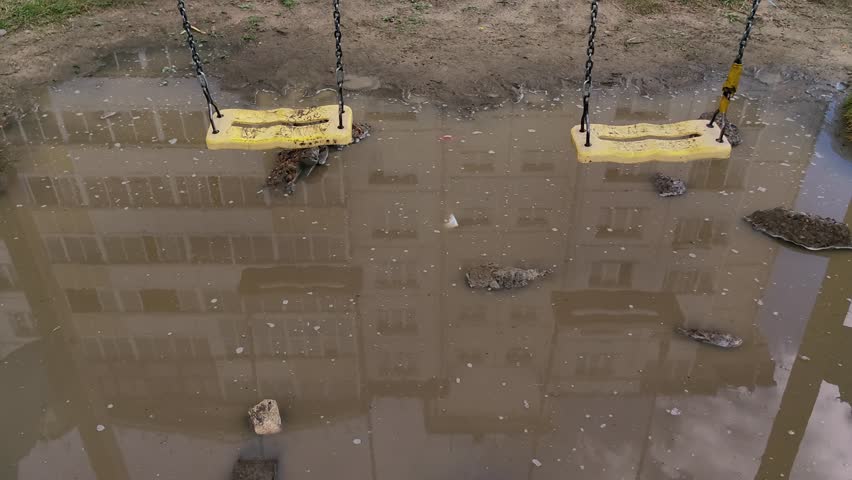 two empty yellow swings swaying gently over flooded playground, with large muddy puddle reflecting residential building after heavy rain, creating somber and lonely atmosphere.