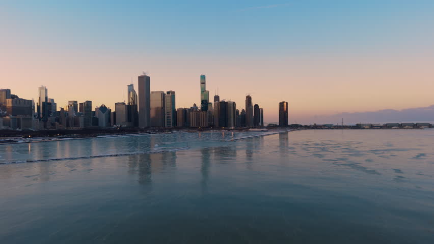 Aerial view of Chicago skyline with Lake Michigan at winter. Thin ice sheets and floating frozen formations cover water surface while warm golden light reflects off the city skyscrapers