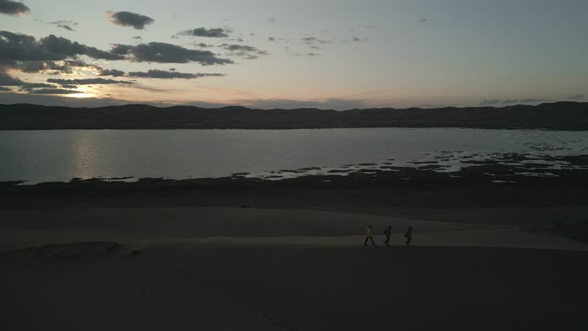 Raw drone footage of group of friends with backpacks and trekking poles walking on sand dune ridge at sunset in Mongolia, wide aerial view of hikers in desert with lake background, unedited.
