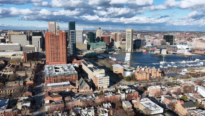 Aerial view of Baltimore townhouses with Inner Harbor and downtown skyline under bright sunlight, showing historic brick architecture, waterfront and urban cityscape of Maryland.