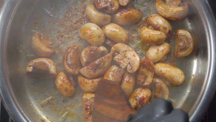 A wooden spatula mixes frying champignon halves with spices in a pan. Top down view. The process of cooking a delicious vegetarian meal or side dish