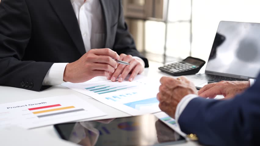 Two businessmen working together, discussing financial data and analyzing product growth charts with a calculator and laptop during a collaborative meeting in a modern corporate office