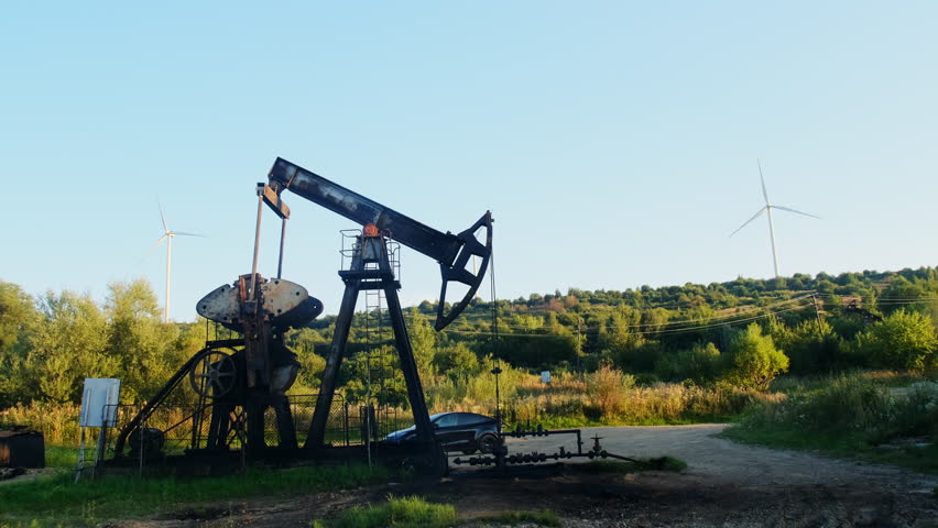 Oil pumpjacks stand on field near gravel track and shrubs. Walking beam swings steadily while head unit sucks releasing during cycle