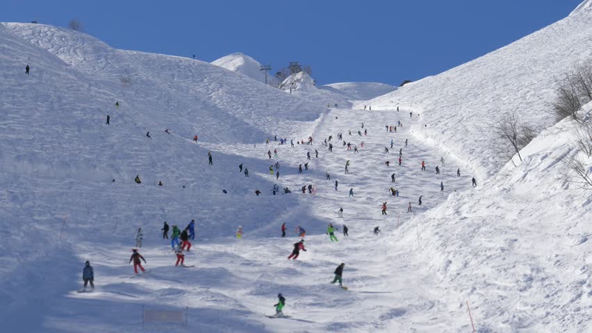 Crowd of skiers descends rugged ski piste. Numerous people skiing and snowboarding down steep, snowy mountain slope at a busy ski resort on a bright sunny day. Winter sports and outdoor activities.