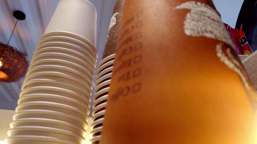 Low-angle view of stacked disposable coffee cups and a syrup bottle on a cafe shelf under warm lighting. Takeaway drinks, barista setup, and coffee shop service concept.