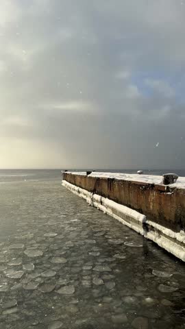 Snow-covered concrete bridge extending into the sea in winter. Cold minimalist seascape with industrial structure surrounded by icy water and calm atmosphere.