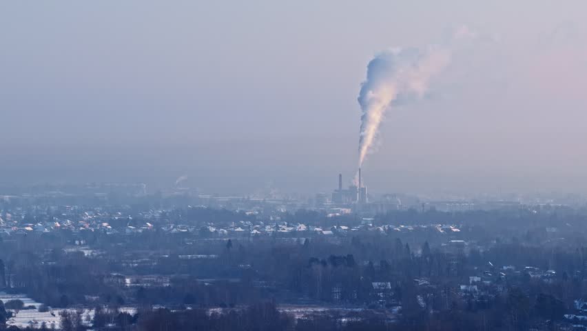 Industrial plume rises through pale sky beyond frozen fields and clustered rooftops