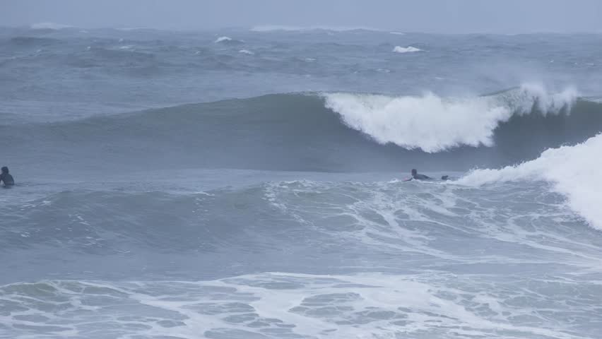 Surfers in wetsuits paddle and ride powerful breaking waves in rough sea conditions. Outdoor adventure and extreme water sport scene.