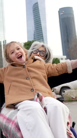 Joyful little girl sitting on the lap of her happy grandmother in a wheelchair, enjoying a walk together in a modern city with skyscrapers, symbolizing family love, care, and quality time