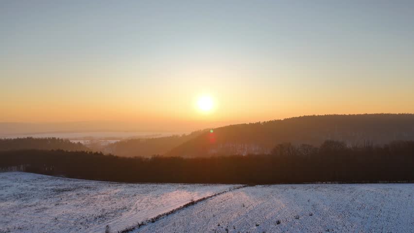 Snowy winter landscape at sunset with rolling hills, distant forest, golden hour light, serene countryside scenery, natural terrain illuminated by warm sunlight. Aerial shot at nature landscape