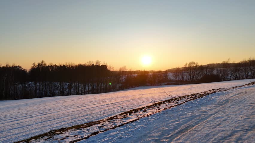 Snowy winter landscape at sunset with rolling hills, distant forest, golden hour light, serene countryside scenery, natural terrain illuminated by warm sunlight. Aerial shot at nature landscape