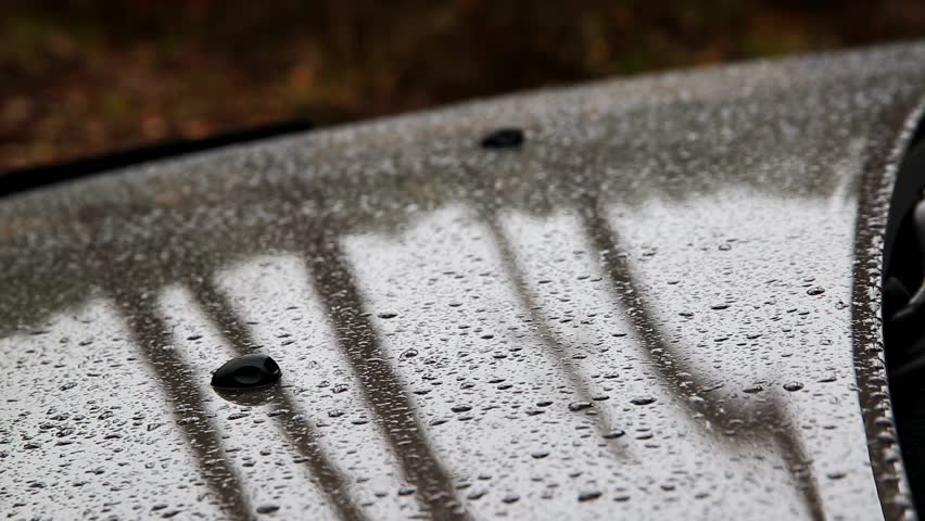 Detailed perspective of numerous rain droplets clinging to polished gray car hood, creating texture and reflecting ambient light. Two black windshield washer jets protrude from surface.