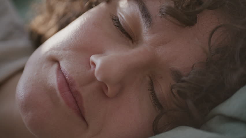 Close up view of curly-haired woman waking up in her bed and gazing away in morning light
