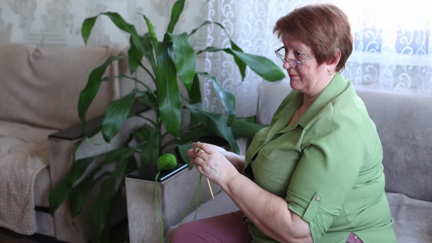 A woman in a green shirt knits with green yarn beside a leafy houseplant, she adjusts her glasses mid stitch, capturing a quiet, focused domestic moment.