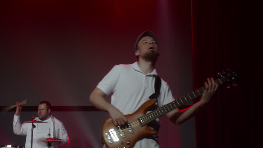 Female singer performing live on stage with raised hands and microphone, supported by guitarist and keyboard player in concert setting.