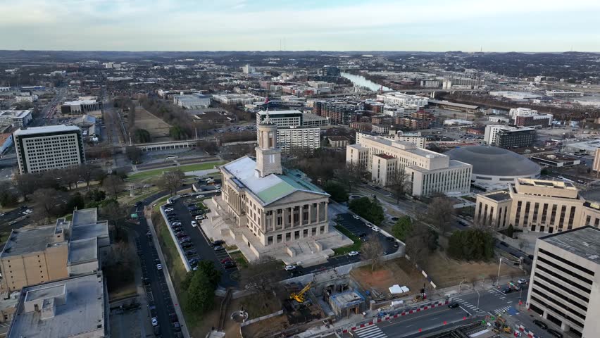 Aerial approaching shot of Tennessee State Capitol in Nashville, showing historic government architecture surrounded by downtown buildings and streets under clear daylight. Waving American flag on top