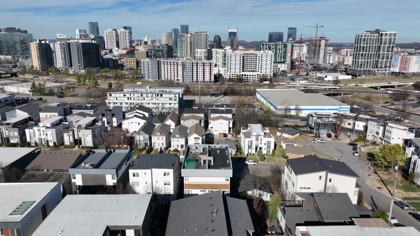 Modern housing in Nashville with contemporary townhomes in foreground and downtown skyline in background, showing urban growth and modern city architecture. Aerial lateral wide shot. Sunny winter day.